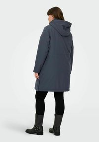 Sieviete stāv ar muguru pret mums, valkājot garu tumši pelēku kapuci ar kapuci, melnas legingas un melnas potītes zābaki ar sprādzēm.