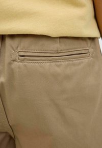 Des pantalons en coton beige au toucher lisse présentent une poche passepoilée horizontale à l'arrière, cousue soigneusement le long du bord.