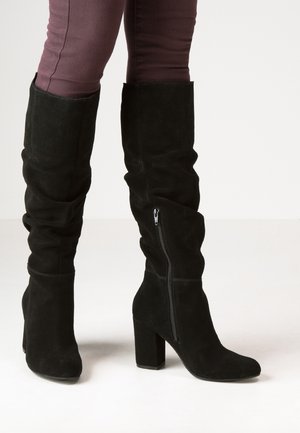Zwarte suède kniehoge laarzen met blokhakken en ritsen aan de zijkant, gedragen met donkerpaarse nauwsluitende broek tegen een effen achtergrond.
