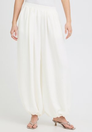 Pantalones blancos de pierna ancha y textura con dobladillos recogidos en los tobillos, usados con sandalias de tacón con estampado de serpiente en una persona de pie.