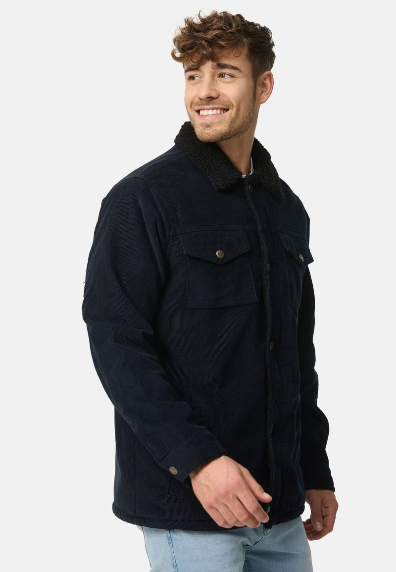 Marineblauwe corduroy jas met een gestructureerde zwarte nepbont kraag, voorknopsluiting en twee borstzakken met gouden accenten.