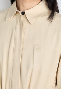 Chemise beige à col, fabriquée en tissu lisse, dotée d'une fermeture par boutons et d'un logo en relief sur le côté gauche. Détails de couture subtils présents.