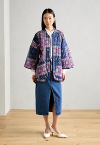 Sieviete, kas valkā ziedotu quilted jaku tumši zilos un rozā toņos, baltu blūzi, zilu midi džinsu svārku ar priekšējo šķēlumu un baltus papēžus, stāv iekštelpās.