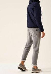 Man som bär en marinblå jacka, grå uppvikta jeans och grå sneakers, står mot en enkel ljus bakgrund, vänd bort.