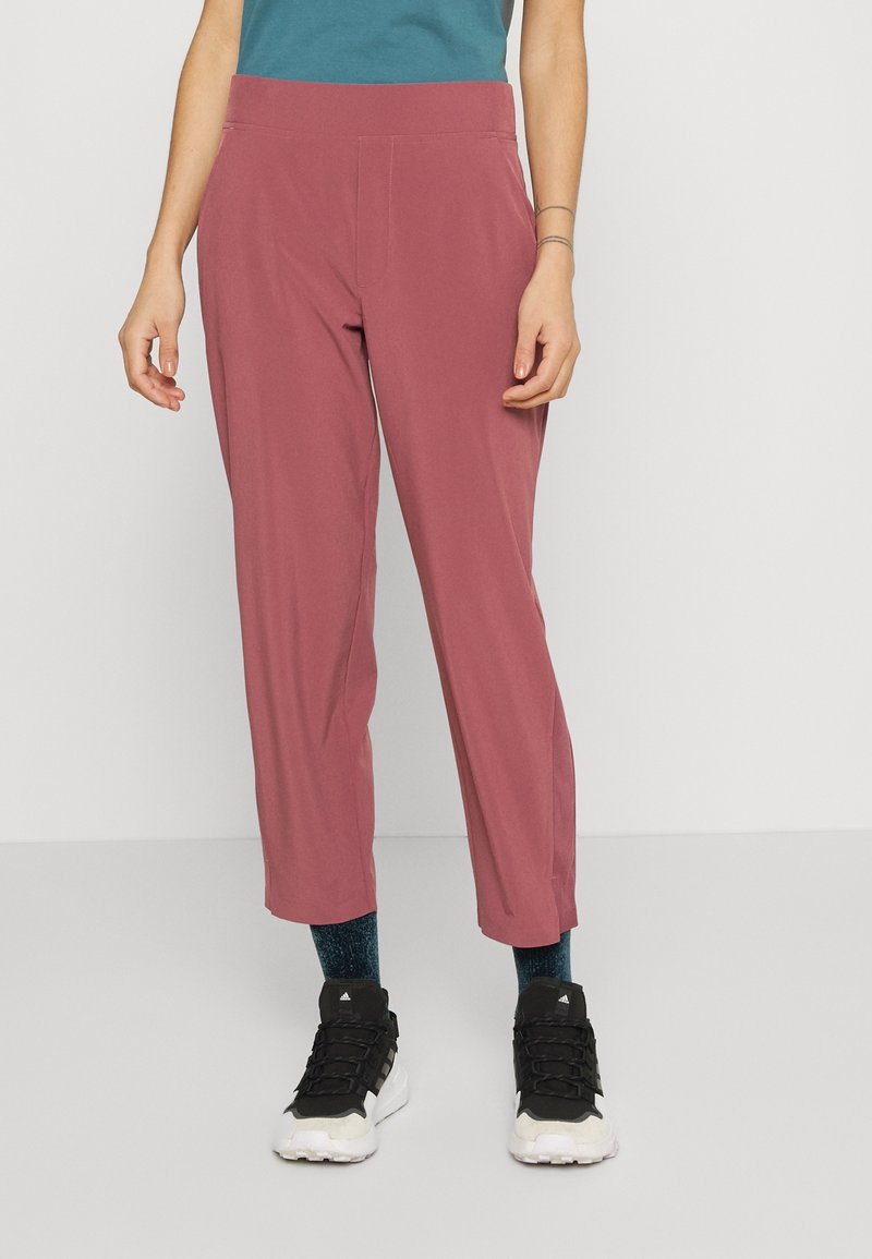 Femme portant un pantalon rose taille haute fuselé, des chaussettes foncées et des chaussures de sport noires et blanches, debout devant un fond uni.