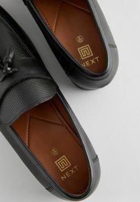 Mocasines de cuero negro con un panel de tela texturizada, presentando un detalle decorativo de nudo y un forro interior marrón. Etiqueta de tamaño visible.
