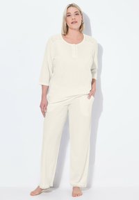 Conjunto de loungewear color beige claro con cuello redondo, mangas de tres cuartos y detalles de ganchillo en los hombros. Los pantalones tienen dobladillos en forma de ondas.