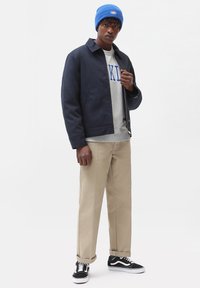 Giacca bomber navy, felpa grigia con scritte blu, pantaloni beige, berretto blu, sneaker bianco e nero. Tessuto liscio, taglio dritto.