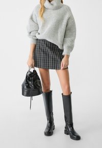 Femme portant un pull gris en maille épaisse, une mini-jupe noire à carreaux, des bottes noires montantes jusqu'aux genoux, tenant un sac seau en cuir noir avec des poignées en chaîne.