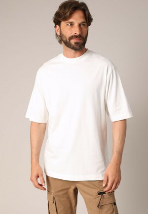 Man met een baard en medium-lang haar, gekleed in een eenvoudig wit oversized t-shirt en lichtbruine cargobroeken, kijkend opzij.