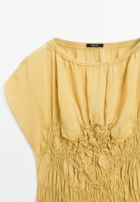 Gelbe, leichte Bluse aus Stoff mit lässiger Passform. Verfügt über gesmokte Details am Kragen und filigrane Muster entlang des Körpers.