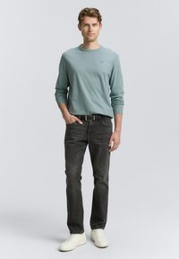 Hellblaues Langarmhemd, graue Jeans und weiße Sneakers. Das Hemd hat einen runden Ausschnitt und dezente Logodetails.