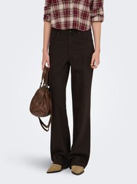 Personne portant un pantalon large marron foncé, une chemise à carreaux, des chaussures pointues couleur beige, tenant un sac à main en cuir marron à côté d'elle.