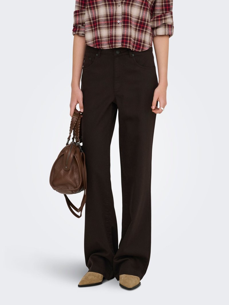 Personne portant un pantalon large marron foncé, une chemise à carreaux, des chaussures pointues couleur beige, tenant un sac à main en cuir marron à côté d'elle.