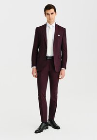 Bordowy garnitur o dopasowanym kroju, z klapami w kształcie litery notch, biała koszula i jasny krawat. Czarne buty dopełniają formalny wygląd.
