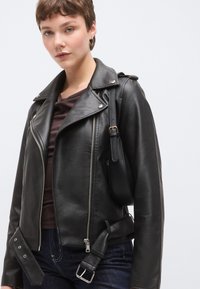 Μαύρο δερμάτινο μπουφάν μοτοσικλέτας με ασημένια φερμουάρ, πλαϊνές αγκράφες και ελαφρώς υφασμένη επιφάνεια, συνδυασμένο με σκουρόχρωμο τοπ και τζιν.