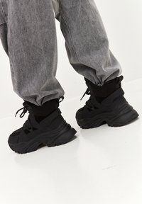 Zapatillas negras con una suela gruesa y texturizada, con una parte superior de malla y un collar acolchado. El diseño incluye correas y cordones ajustables.