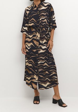 Femme portant une robe midi boutonnée avec des motifs ondulés noirs, marron et beige, associée à des chaussures noires à talons blocs ouvertes à l'avant.