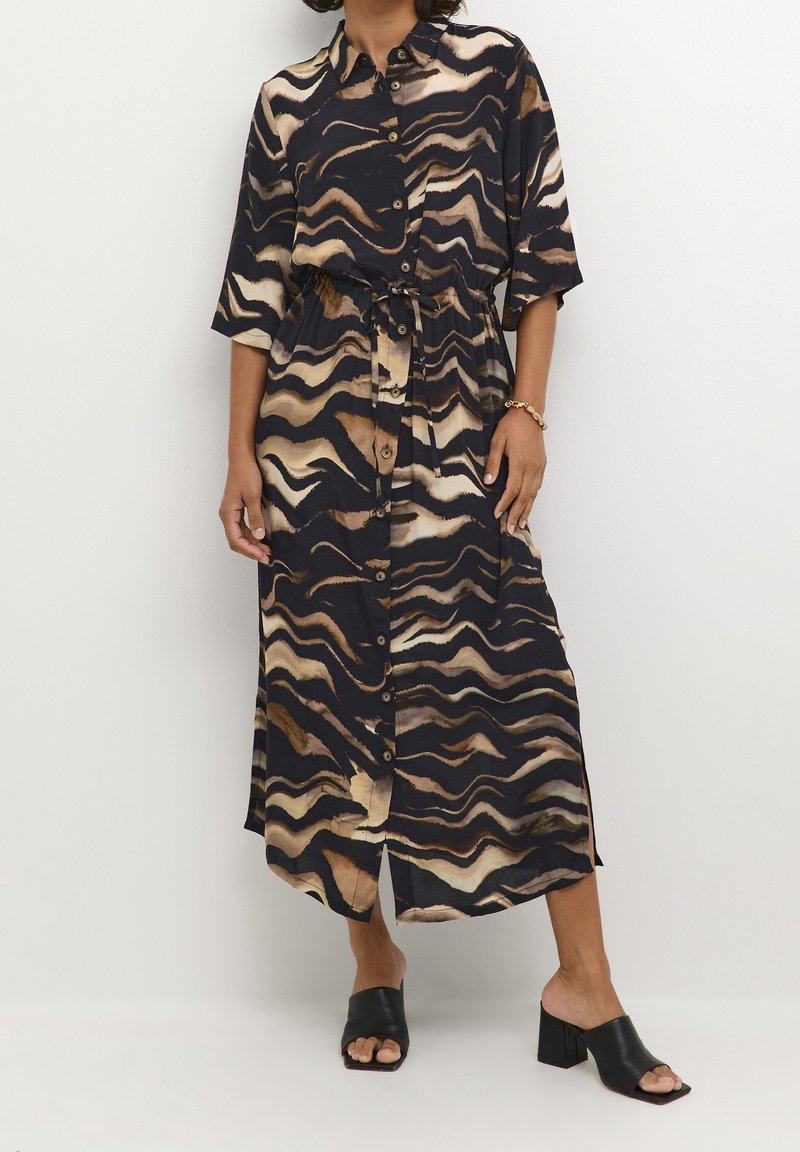 Femme portant une robe midi boutonnée avec des motifs ondulés noirs, marron et beige, associée à des chaussures noires à talons blocs ouvertes à l'avant.