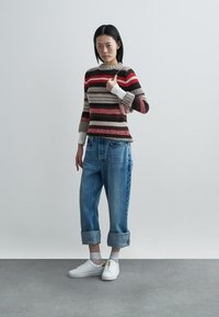 Knitted sweater with horizontal stripes in beige, red, and brown. Paired with cuffed light blue jeans and white sneakers featuring gold zippers.