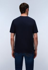 Mann mit kurzen braunen Haaren von hinten gesehen, trägt ein schlichtes dunkelblaues T-Shirt und blaue Jeans vor grauem Hintergrund.