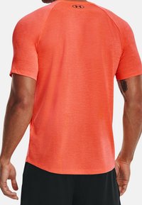 Camiseta atlética de manga corta en un vibrante color naranja, fabricada con un tejido texturizado. Presenta un dobladillo redondeado y un pequeño logo en la parte posterior del cuello.