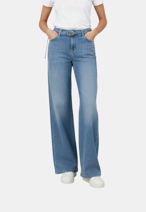 Modello che indossa jeans a gamba larga azzurri chiari con cintura e sneaker bianche, mani nelle tasche, abbinato a una maglietta bianca a maniche corte.