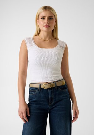 Kobieta w białej, teksturowanej, bezrękawnikowej bluzce, ciemnoniebieskich jeansach, beżowym pasku, złotych kolczykach kołach i złotym naszyjniku na łańcuszku, stojąca na białym tle.