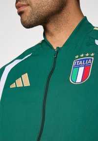 Zelená športová bunda so zipsom, biele pruhy, zlaté logo Adidas, nášivka Taliansko s štyrmi hviezdami a trojfarebným emblémom.