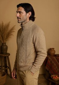 Pull en tricot beige avec un col montant, présentant des motifs en maille torsadée et des poignets côtelés. Associé à un pantalon beige sur un fond chaud et neutre.