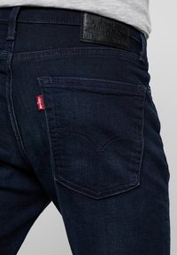 Tmavě modré džínsy z denimu s černou koženou nášivkou na pasu. Mají charakteristickou zadní kapsu s detailním prošitím ve tvaru srdce.