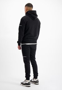 Zwarte hoodie met geribde manchetten, gecombineerd met bijpassende joggingbroek. Opvallende details zijn het merk op de mouw en de zijkant van de broek.