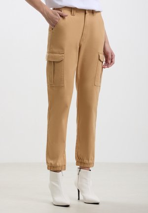 Frau trägt beige Cargohosen mit elastischen Bündchen und weiße hochhackige Stiefeletten vor einfachem Hintergrund.