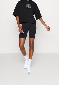 Svart oversized hoodie med DKNY Sport-logotyp, kombinerad med svarta cykelshorts med sidografik; vita sneakers med rosa detaljer.