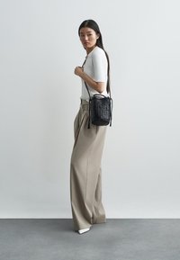 Μαύρη πλεκτή τσάντα με κούμπωμα κορδονιού, συνδυασμένη με εφαρμοστό άσπρο ζακέτα ριμπ και μπεζ φαρδιά παντελόνα. Λείο υλικό και μινιμαλιστικός σχεδιασμός.