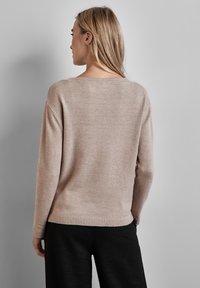 Beige gestrickter Pullover mit lockerer Passform und weitem Ausschnitt, mit gerippten Bündchen und Saum, von hinten vor einem grauen Hintergrund betrachtet.