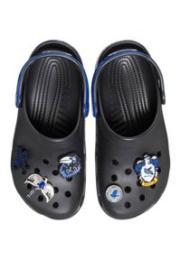 Black clogs with blue straps, featuring decorative charms: a raven, a silver wing, and a blue circular badge. Textured interior and vent holes.