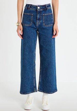 Vrouw draagt wijde blauwe spijkerbroek met grote voorzakken, witte sneakers, een wit topje en een gouden armband om haar linkerpols.