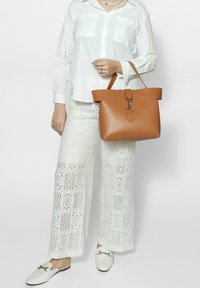 Borsa a tracolla in pelle color cuoio con forma strutturata, texture liscia e chiusura a scatto. Indossata con una camicia bianca e pantaloni con motivo a pizzo.