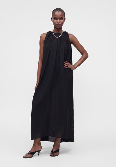 Femme portant une robe longue noire sans manches, des sandales à talons noires et un collier simple, debout avec une main sur la hanche.
