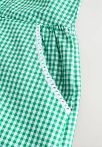 Vihreä ja valkoinen ruudullinen kangas, jossa on kootut saumat ja koristeellinen valkoinen pitsireunus sivutaskun ympärillä. Sujuva pinta.