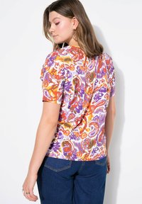 Korte mouwen, aansluitend shirt in witte stof met een kleurrijk paisleypatroon in paarse, oranje en rode tinten. Zachte textuur, ronde hals.