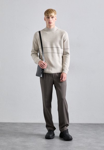 Beige stickad tröja med ribbad krage, mörka horisontella ränder, grå byxor och svart axelremsväska, bärs med svarta sneakers.