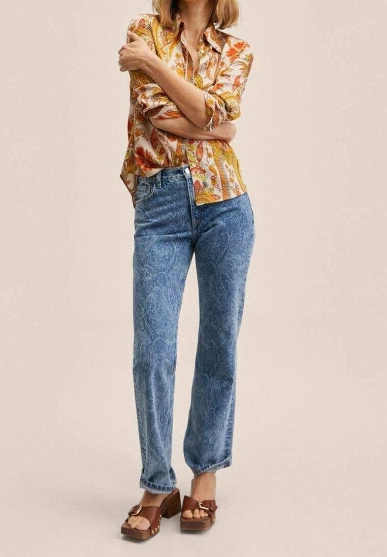 Femme portant un chemisier orange et jaune à motifs, un jean bleu taille haute et des sandales marron à talons, debout les bras croisés.