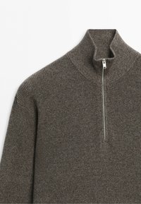 Brun stickad tröja med hög krage och kort silverfärgad dragkedja framtill på en enfärgad vit bakgrund.