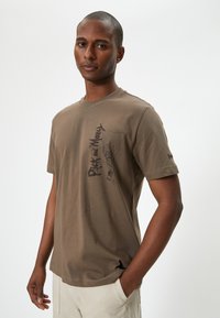 Brun bomulds T-shirt med korte ærmer, der har trykt en sort grafisk illustration af en figur og tekst på forsiden, rund halsudskæring.