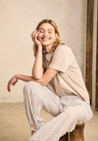 Mosolygó nő csukott szemmel, bézs pólót és fehér vékony csíkos nadrágot viselve, egy fakorben ülve, egyszínű fal előtt.