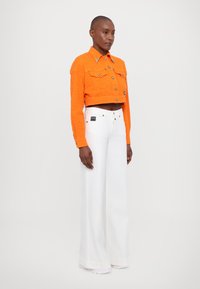 Giacca di jeans arancione a trama cropped con bottoni dorati e due tasche frontali, abbinata a jeans bianchi a gamba larga con etichetta nera.