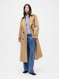 Manteau trench beige avec des boutons noirs, grands revers, et une taille ceinturée, associé à une chemise rayée bleu et blanc et un jean bleu.