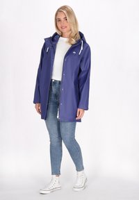 Violetinė nepralaidi vandeniui striukė su kapišonu, priekyje užtrauktuku ir su raišteliais akcentais, derinama su mėlynomis liesomis kelnėmis ir baltomis sportinėmis bateliais.